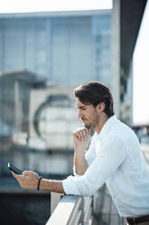 Businessman with hand on chin using mobile phone leaning on railing - GUSF07182