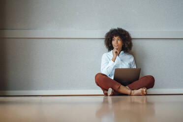 Thoughtful businesswoman with laptop sitting on floor at work place - JOSEF07383