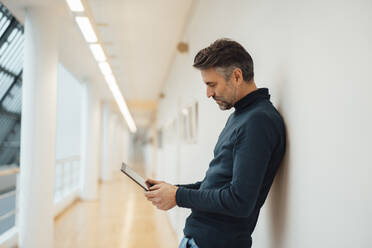 Businessman using tablet computer leaning on wall in corridor - JOSEF07403