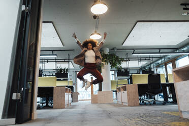 Cheerful businesswoman with hands raised jumping in office - JOSEF07411