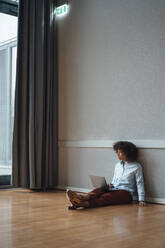 Thoughtful businesswoman with laptop sitting on floor in office - JOSEF07440