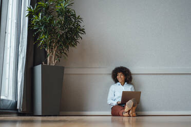 Geschäftsfrau mit Laptop träumt vor einer Wand im Büro - JOSEF07444