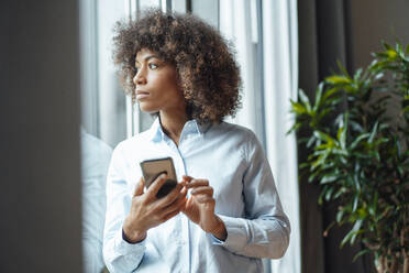 Businesswoman with smart phone looking through window in office - JOSEF07446