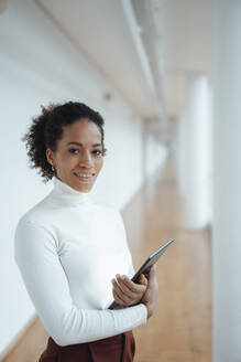 Lächelnde berufstätige Frau mit Tablet-PC im Bürokorridor - JOSEF07523