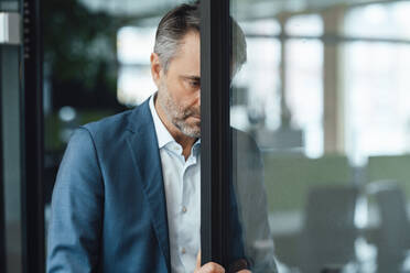 Businessman leaning head on glass door at work place - JOSEF07530