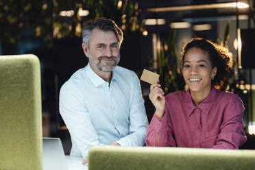 Happy businesswoman with credit card sitting by colleague at work place - JOSEF07546