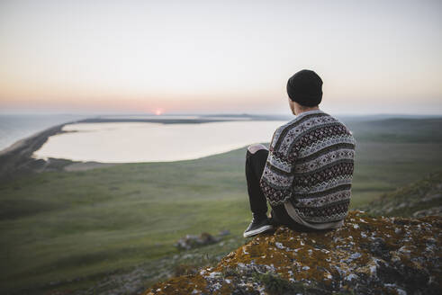 Junger Mann im gestreiften Pullover sitzt bei Sonnenuntergang auf einem Hügel - TETF00769