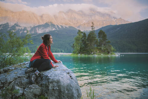 Deutschland, Bayern, Eibsee, Junge Frau sitzt auf einem Felsen und blickt auf eine malerische Landschaft am Eibsee in den bayerischen Alpen - TETF00771