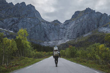 Norway, Lofoten Islands, Backpacker walking down road in mountain landscape - TETF00773