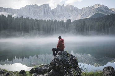 Switzerland, Young man sitting at Lago di Carezza in Dolomite Alps at dawn - TETF00778