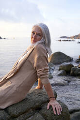 Frau mit grauem Haar sitzt auf einem Felsen am Strand - VEGF05384