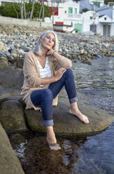 Nachdenkliche Frau mit grauem Haar sitzt auf einem Felsen am Strand - VEGF05387