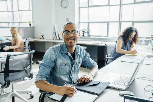 Smiling man using note pad by laptop in office - TETF00873