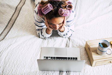 Frau mit Lockenwicklern auf dem Bett liegend und mit Blick auf den Laptop - TETF00888