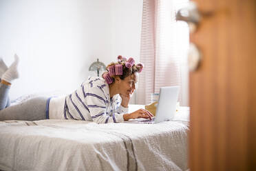 Frau mit Lockenwicklern auf dem Bett liegend und mit Laptop - TETF00889