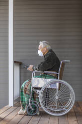 Senior man in wheelchair wearing protective mask to prevent coronavirus transmission on porch - TETF00962