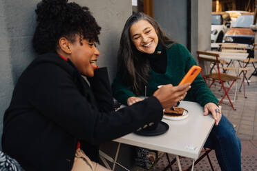 Happy African American female with curly hair sitting at table with dessert and showing cellphone to mature Hispanic female in outdoor cafe - ADSF33796