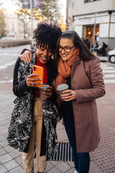 Cheerful adult black lady showing cellphone screen to smiling female friend while standing together on city street with cups of coffee - ADSF33798