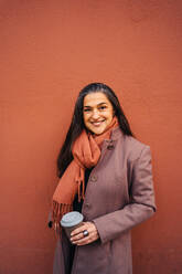 Lächelnde ethnische erwachsene Frau in Mantel und Turnschuhen steht in der Nähe der roten Wand auf der Straße mit Kaffee zum Mitnehmen - ADSF33799