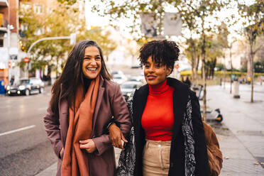 Cheerful lady holding arms black female friend in warm clothes while walking together on street pavement looking away - ADSF33803