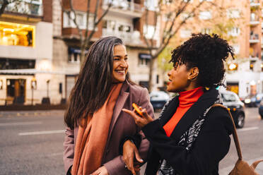 Fröhliche Dame hält Arme schwarze weibliche Freundin in warmer Kleidung beim Gehen zusammen auf der Straße Bürgersteig Blick auf einander - ADSF33804