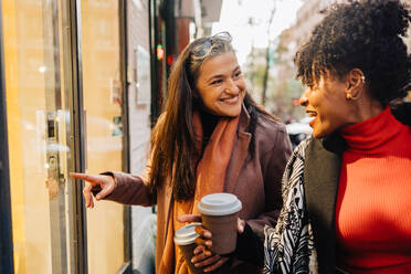 Positive Hispanic lady pointing at glass wall of shop to black female friend on autumn street with takeaway coffee - ADSF33805