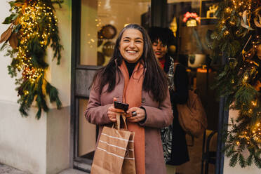 Glückliche Frauen, die nach dem Weihnachtseinkauf mit ihren Einkäufen aus einem dekorierten Geschäft gehen - ADSF33806