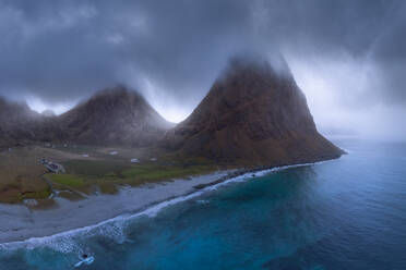 Plätscherndes Meer wäscht grasbewachsenen Ufer mit Gebäude und hohe felsige Klippen gegen nebligen Himmel in Lofoten Inseln in Norwegen gelegen - ADSF33816