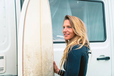 Positiver weiblicher Surfer mit wasserdichtem Neoprenanzug, der mit einem Surfbrett in der Nähe eines Anhängers in der Natur steht und sich auf das Surfen im Sommer vorbereitet - ADSF33843