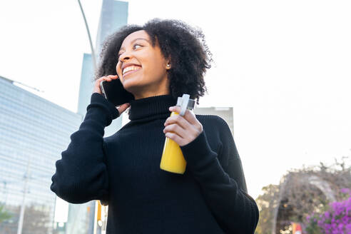 Positive African American female with orange juice looking up while on a phone call on smartphone standing near blooming bushes with purple flowers - ADSF33859