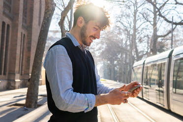 Seitenansicht eines lächelnden, bärtigen hispanischen männlichen Fahrgastes, der auf einem Bahnsteig an einem sonnigen Sommertag auf ein Smartphone wartet - ADSF33860