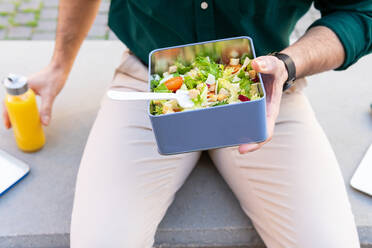 Anonymer männlicher Unternehmer mit einer Lunchbox mit frischem Salat und einem Netbook, das während der Mittagspause auf einer Steinplatte steht - ADSF33863