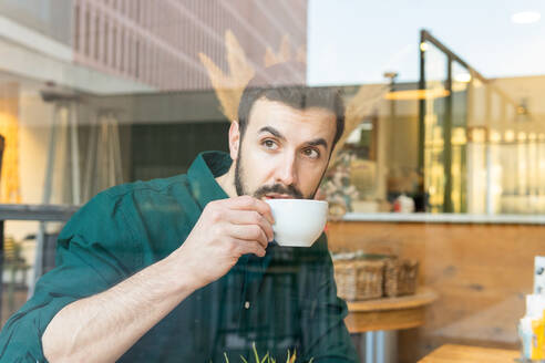 Durch Fenster hindurch nachdenklicher bärtiger männlicher Unternehmer, der heißen Kaffee trinkt, während er mit einem Stück Kuchen in einer hellen modernen Cafeteria während der Mittagspause am Tisch sitzt - ADSF33864