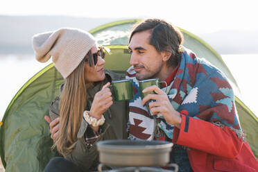 Couple of explorers in outerwear sitting in tent at campsite and enjoying hot drink on shore of lake in mountains in autumn - ADSF33866