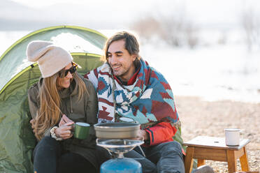 Couple of explorers in outerwear sitting in tent at campsite and enjoying hot drink on shore of lake in mountains in autumn - ADSF33867
