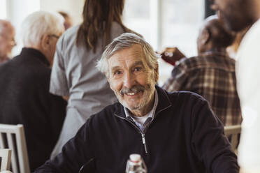Porträt eines lächelnden älteren Mannes mit Pfleger und Freunden im Hintergrund in einem Pflegeheim - MASF28973