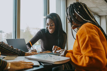 Cheerful female friends studying together while sitting at table in college cafeteria - MASF29085