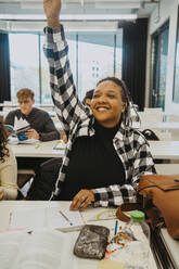 Fröhliche Schülerin hebt die Hand, während sie am Schreibtisch im Klassenzimmer sitzt - MASF29091