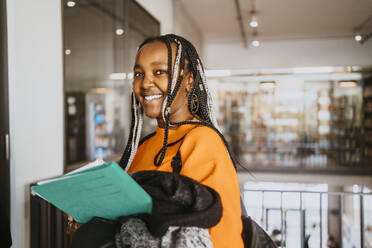 Portrait of smiling braided female student holding jacket and file in university - MASF29153