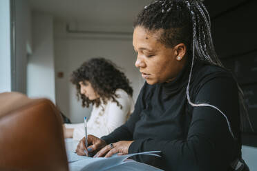 Frau mit geflochtenen Haaren, die in ein Buch schreibt, während sie neben einem Freund in der Universität sitzt - MASF29158