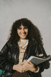 Portrait of smiling young woman wearing leather jacket holding book leaning on gray wall - MASF29192