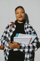 Portrait of smiling mature woman holding book standing against wall on sunny day - MASF29198