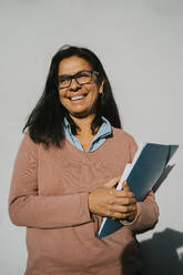 Glückliche reife Frau mit Buch und Akte, die an einer grauen Wand in der Universität an einem sonnigen Tag steht - MASF29200