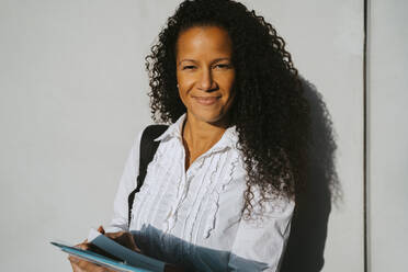 Portrait of smiling woman with curly hair holding book against wall at college campus - MASF29201