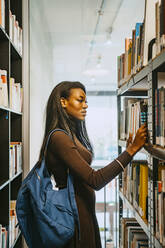Seitenansicht einer jungen Frau mit Rucksack, die in einer Bibliothek ein Buch sucht - MASF29226