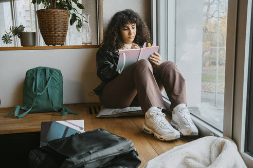 Junge Frau schreibt in voller Länge in ein Buch, während sie in der Universität am Fenster sitzt - MASF29251