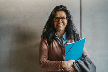Lächelnde reife Frau mit Büchern und Akten an der grauen Wand einer Universität - MASF29259
