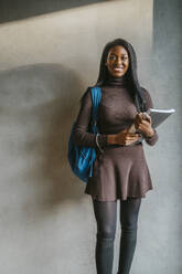 Full length portrait of smiling young woman holding book standing with backpack against gray wall - MASF29260