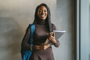 Portrait of cheerful young woman holding book standing with backpack against gray wall - MASF29262