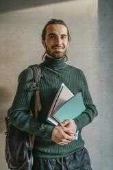 Portrait of smiling young male adult student holding book standing in front of gray wall at college - MASF29264